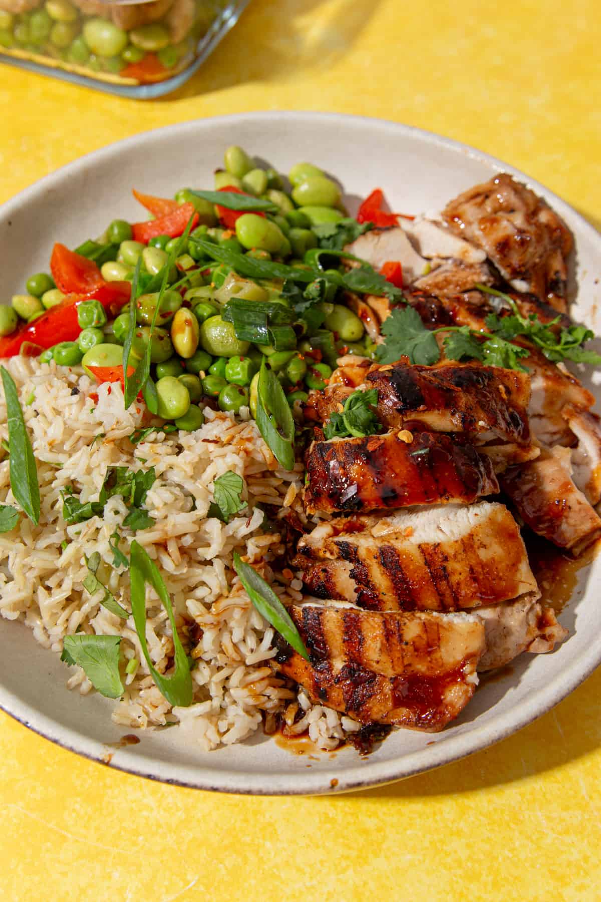 Browned and sliced chicken in a bowl with rice, edamame beans, peas, peppers and spring onion and coriander topping.