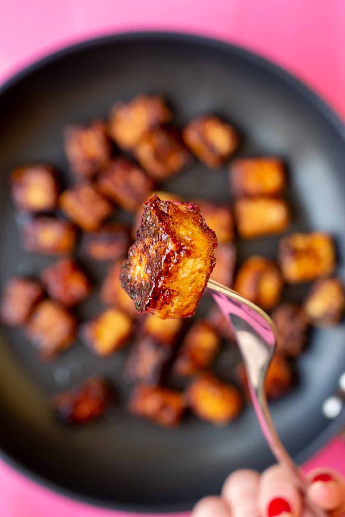 Close up shot of a piece on crispy tofu on a fork with the pan full of the tofu faded in the background.