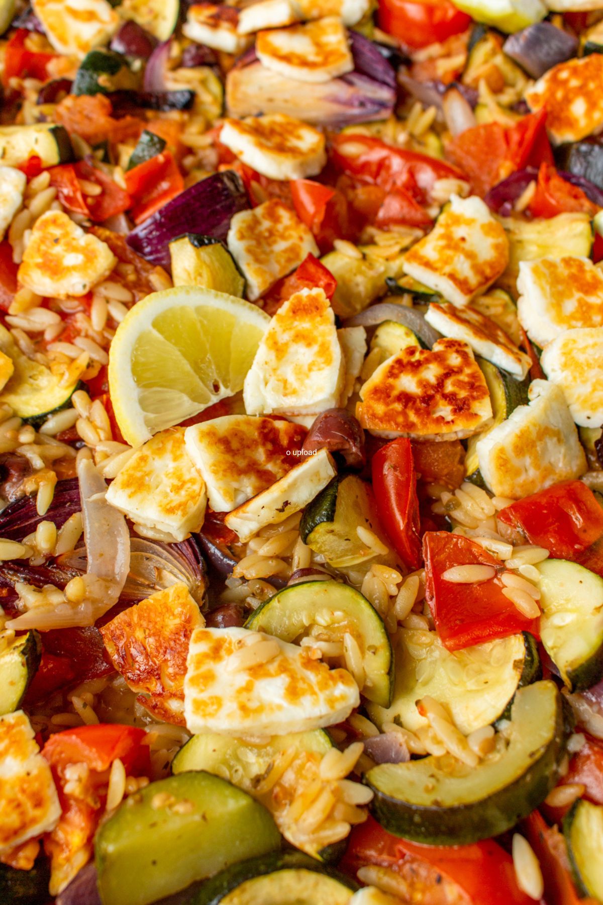 Close up of tray bake with Vegetables, Halloumi and Baked Orzo with red onions, tomatoes and courgettes with lemon wedges.
