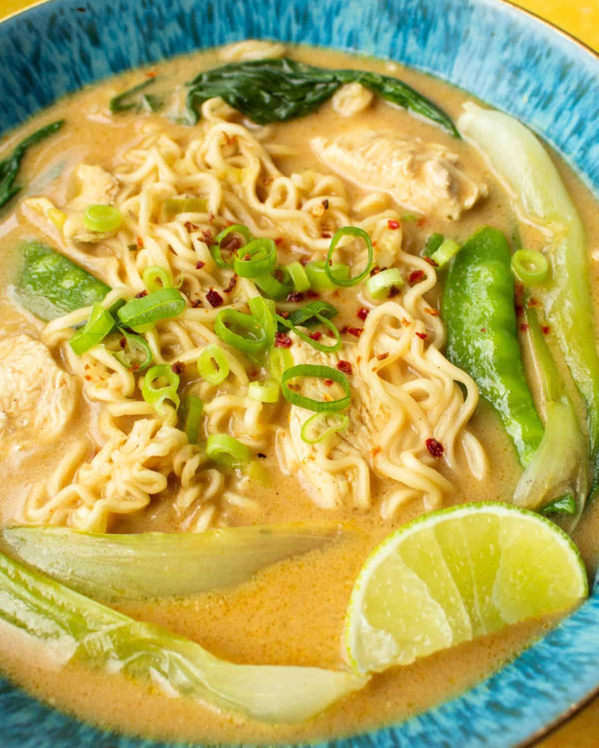 Chicken ramen in a blue bowl with mange tout, noodles, pack choi and sliced spring onions on top with a wedge of lime.