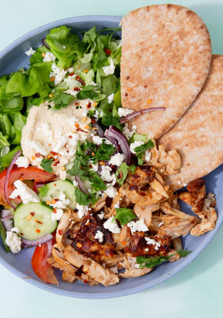 Baked and shressd chicken with pitta bread and salad, with hummus and feta in a blue bowl.