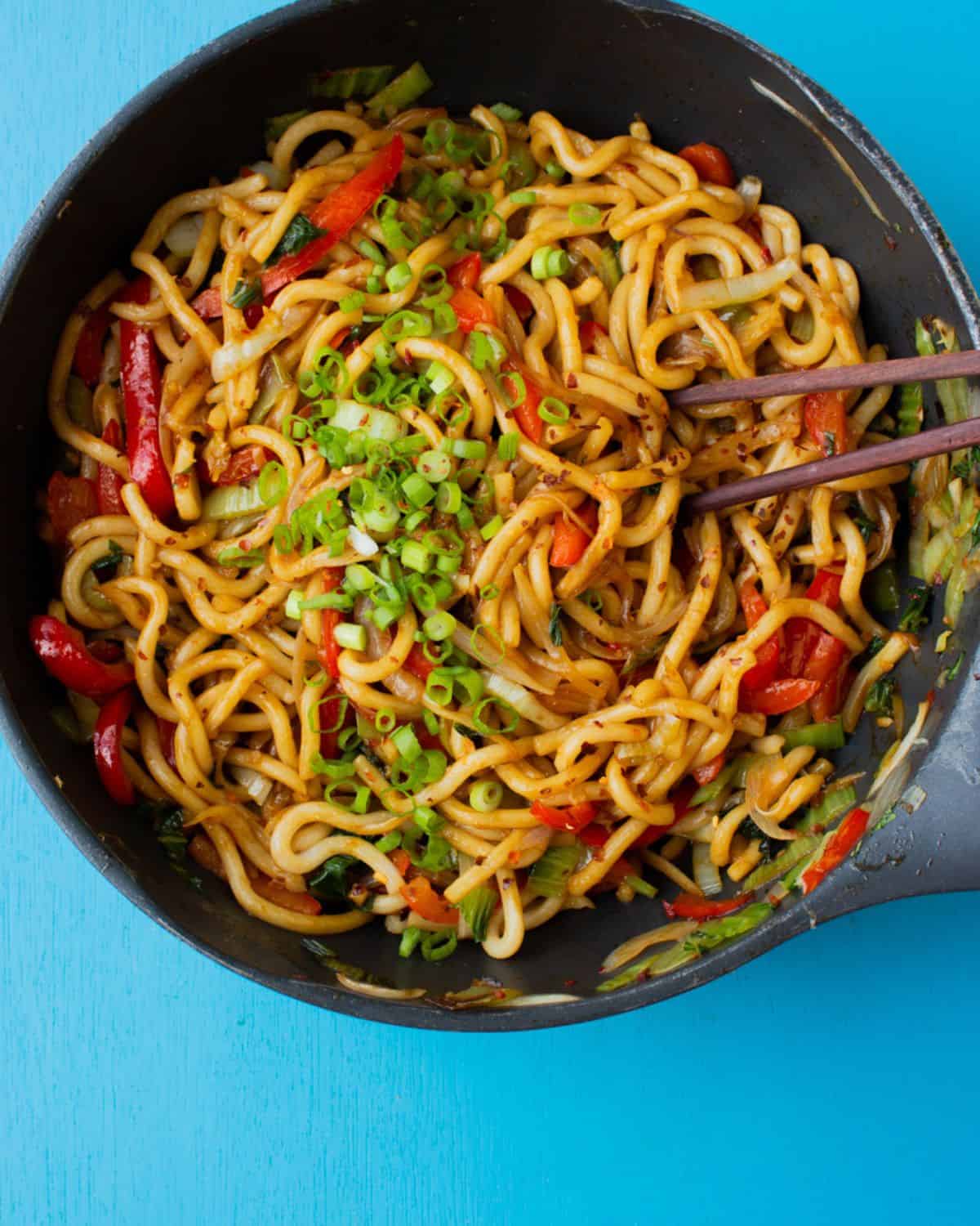 Udon noodles  in a large pan with sliced spring onions, red peppers and oil with chop sticks.