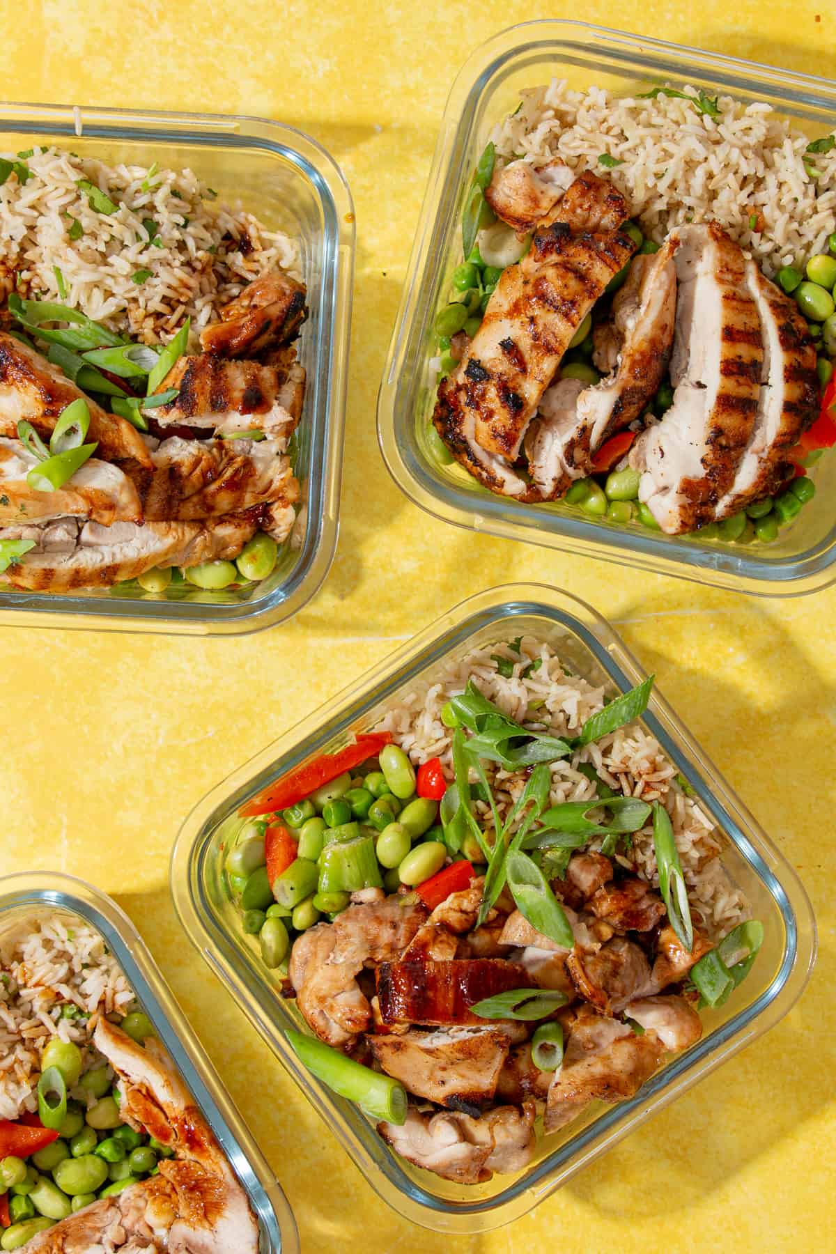 4 square, glass meal prep containers filled with browned and sliced chicken with rice, edamame beans, peas, peppers and spring onion and coriander topping.