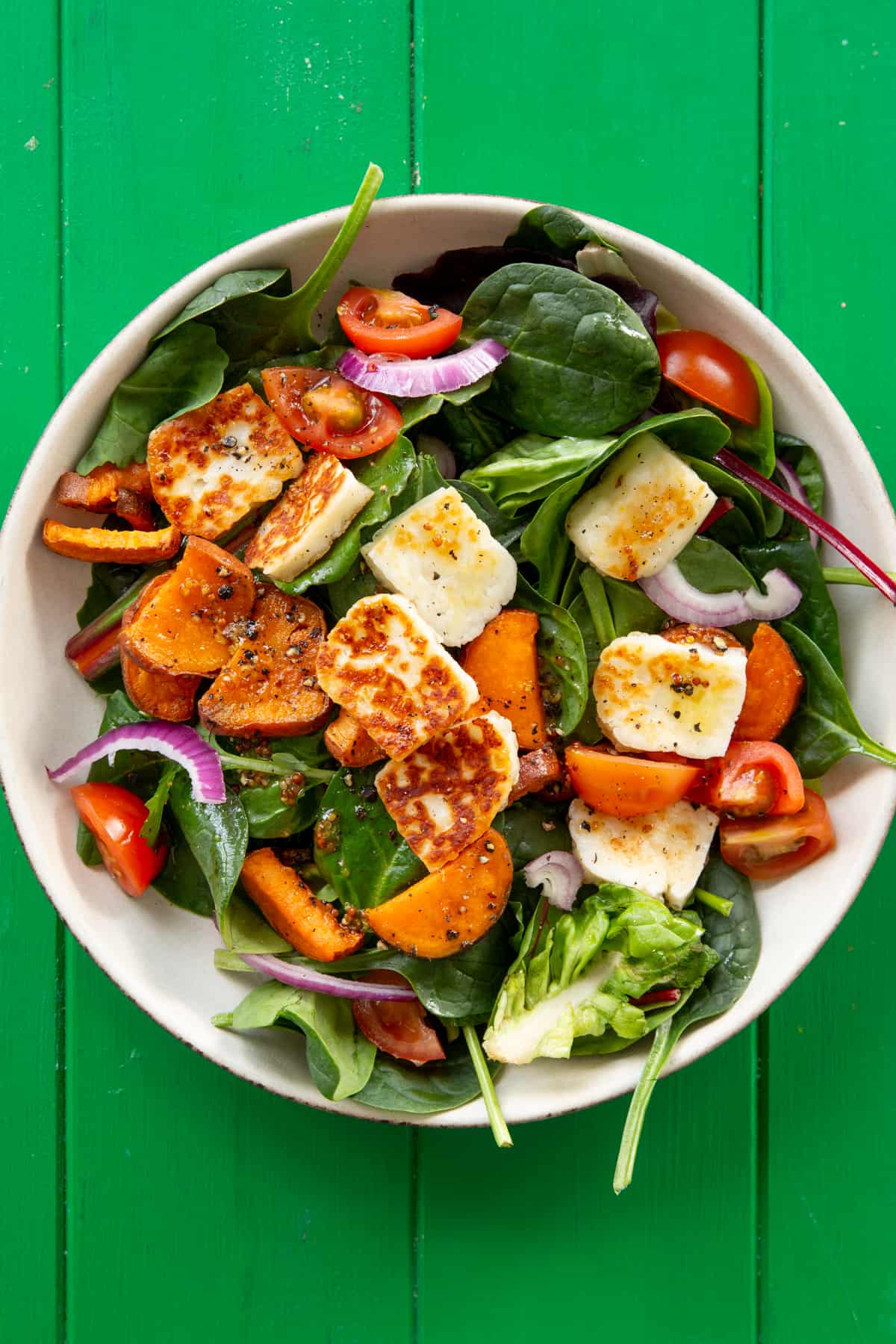 Overhead shot of salad in a bowl with golden browned halloumi, butternut squash, red onion and tomatoes on a green background.