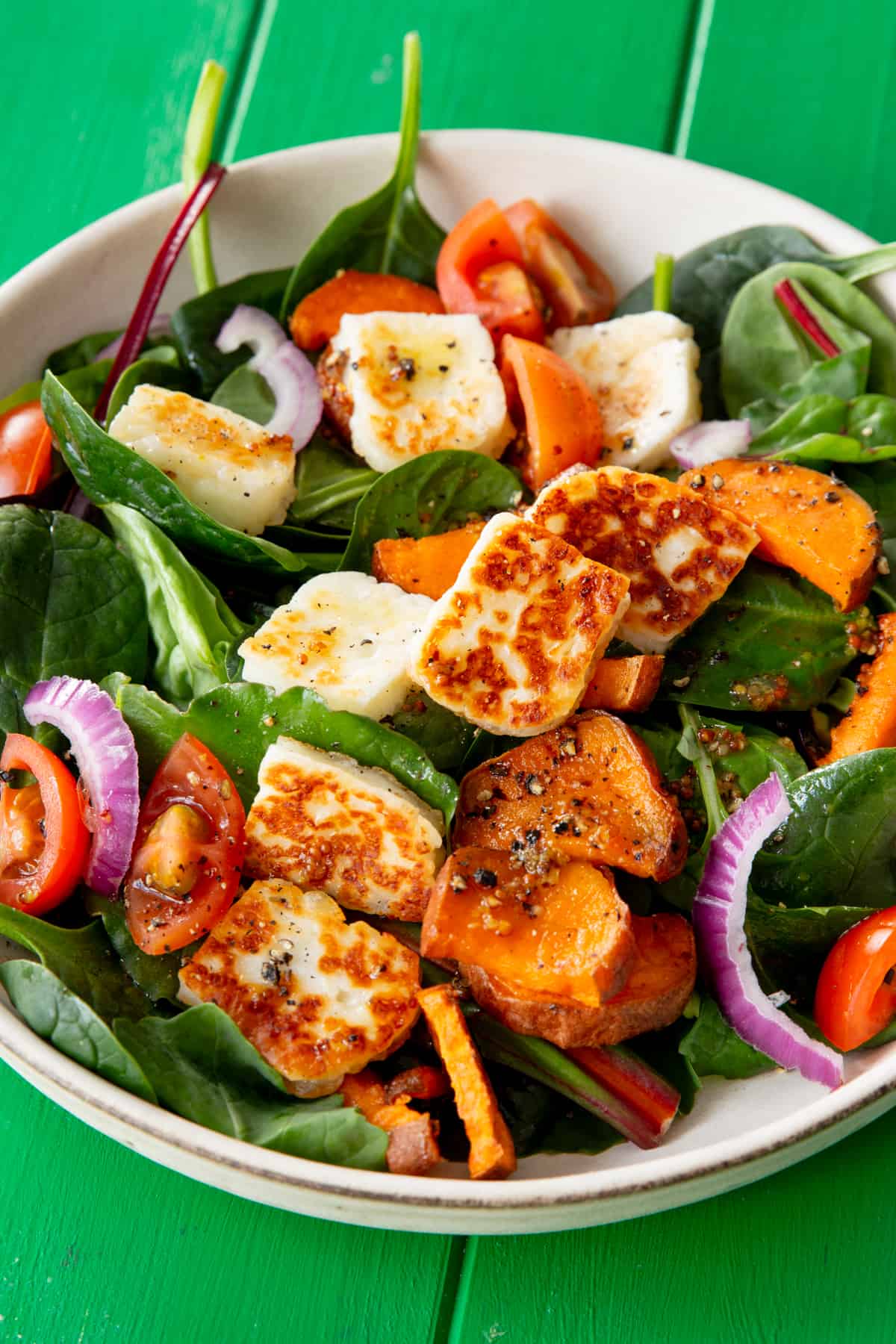 Close up of salad in a bowl with golden browned halloumi, butternut squash, red onion and tomatoes on a green background.