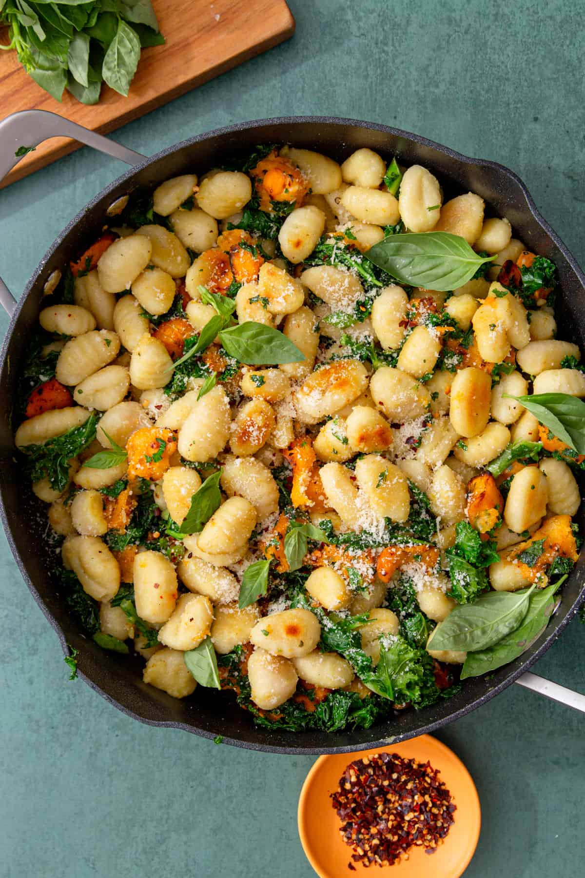 Browned gnocchi in a frying pan with butternut squash and grated  parmesan with kale next to a chopping board with basil leaves and below pan a bowl of chilli flakes.