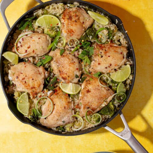 Golden browned chicken thighs in a pan on rice with lime wedges, mint and sliced spring onions on a yellow background.