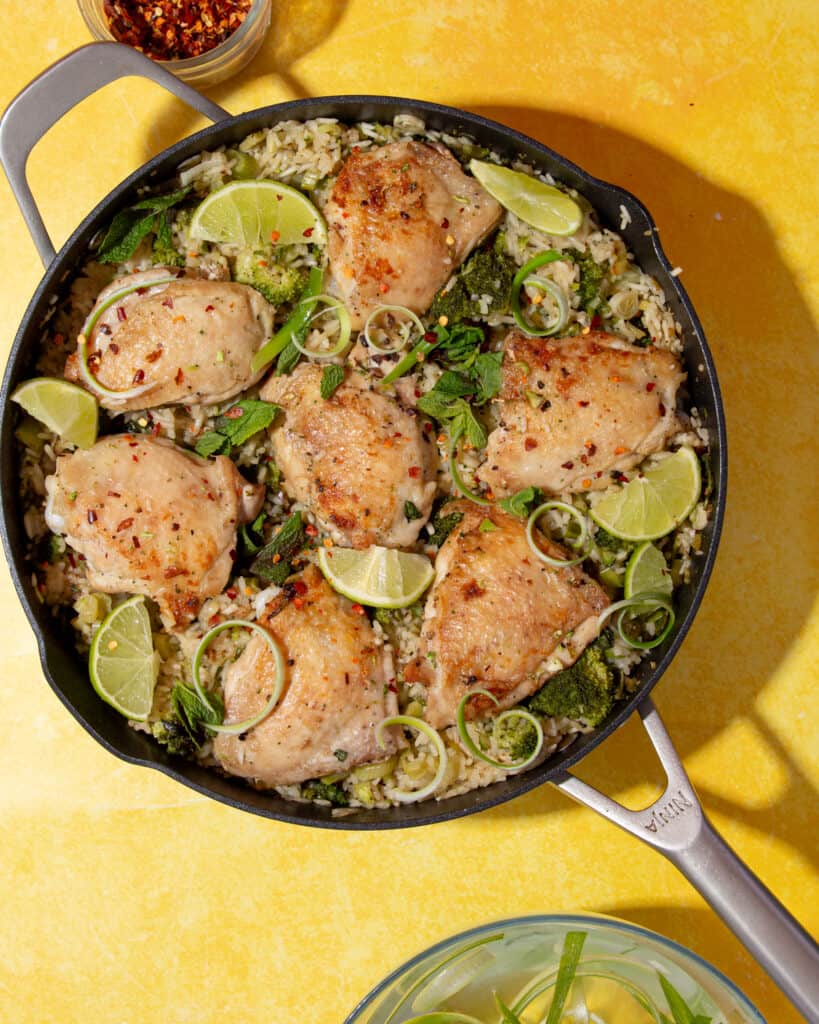 Golden browned chicken thighs in a pan on rice with lime wedges, mint and sliced spring onions on a yellow background.