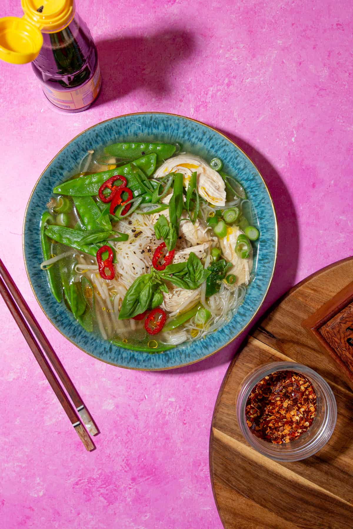 Overhead shot of chicken soup with noodles, mange tout, chillies and spring onions in a blue bowl on a pink background with chop sticks and chilli flakes by dish.