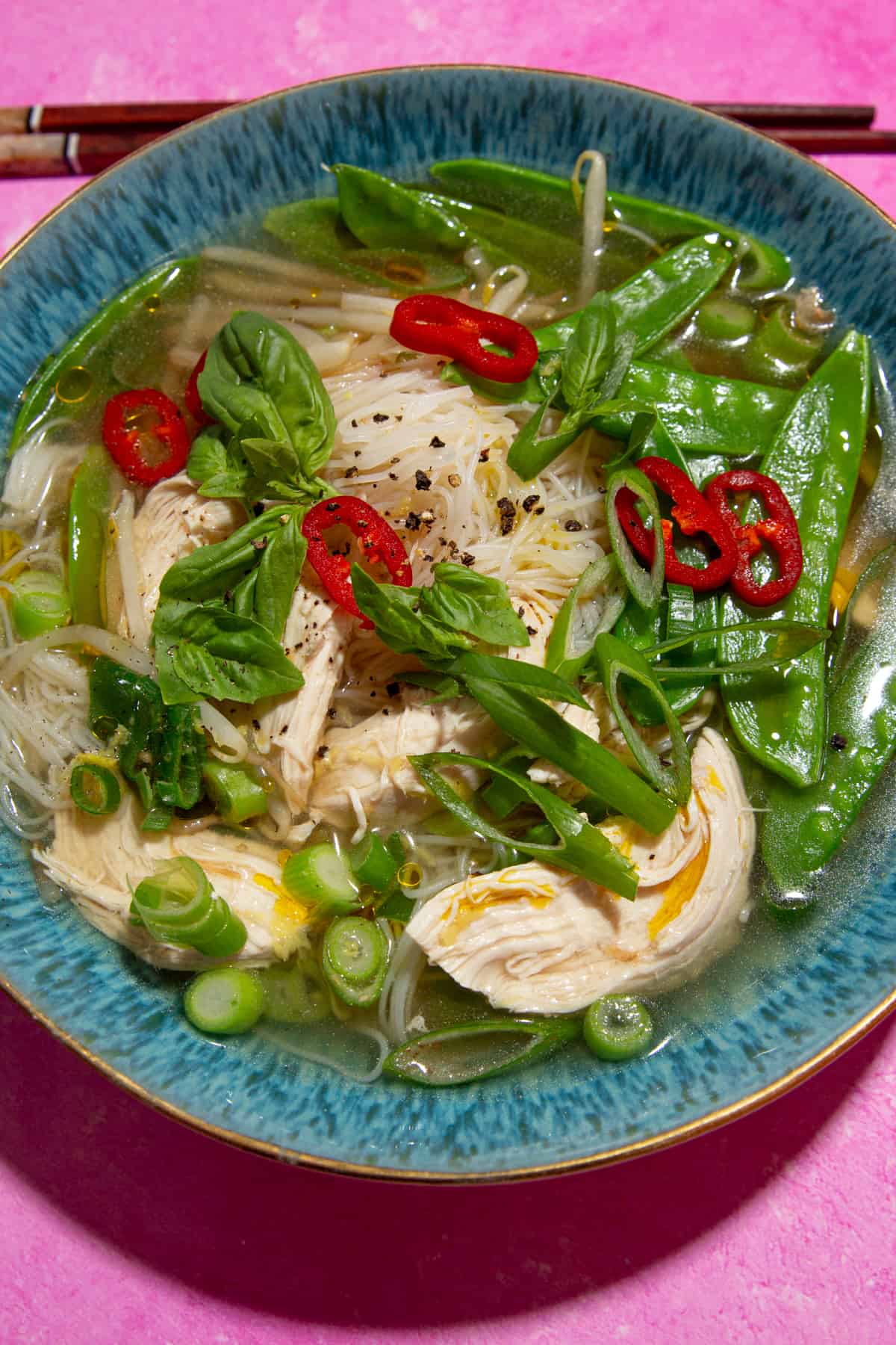 Close up shot of chicken soup with noodles, mange tout, chillies and spring onions in a blue bowl on a pink background with chop sticks laying behing the dish.