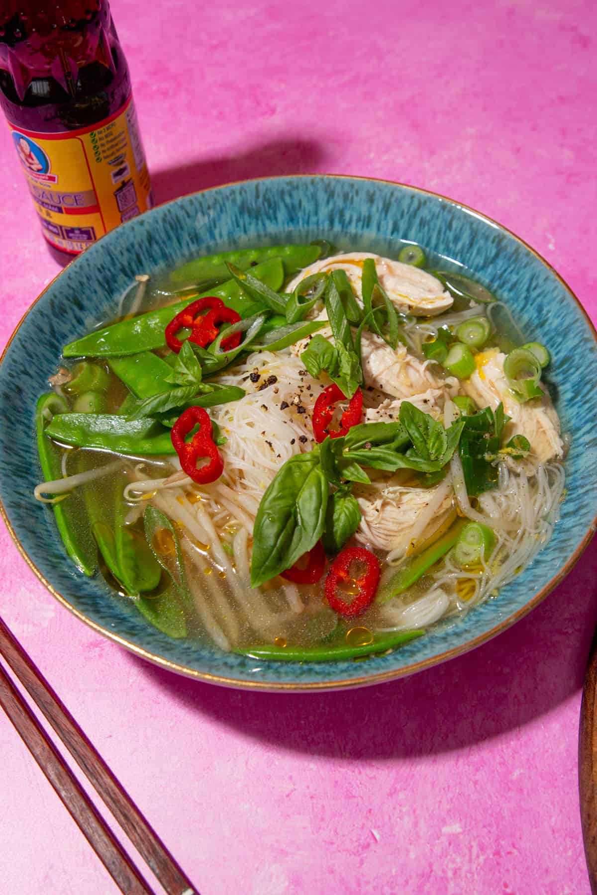 Chicken soup with noodles, mange tout, chillies and spring onions in a blue bowl on a pink background with a bottle of fish sauce behind the dish and chop sticks towards the front.