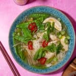 Chicken noodle soup with mange tout, chillies and spring onions in a blue bowl on a pink background with chopsticks. by side of dish.
