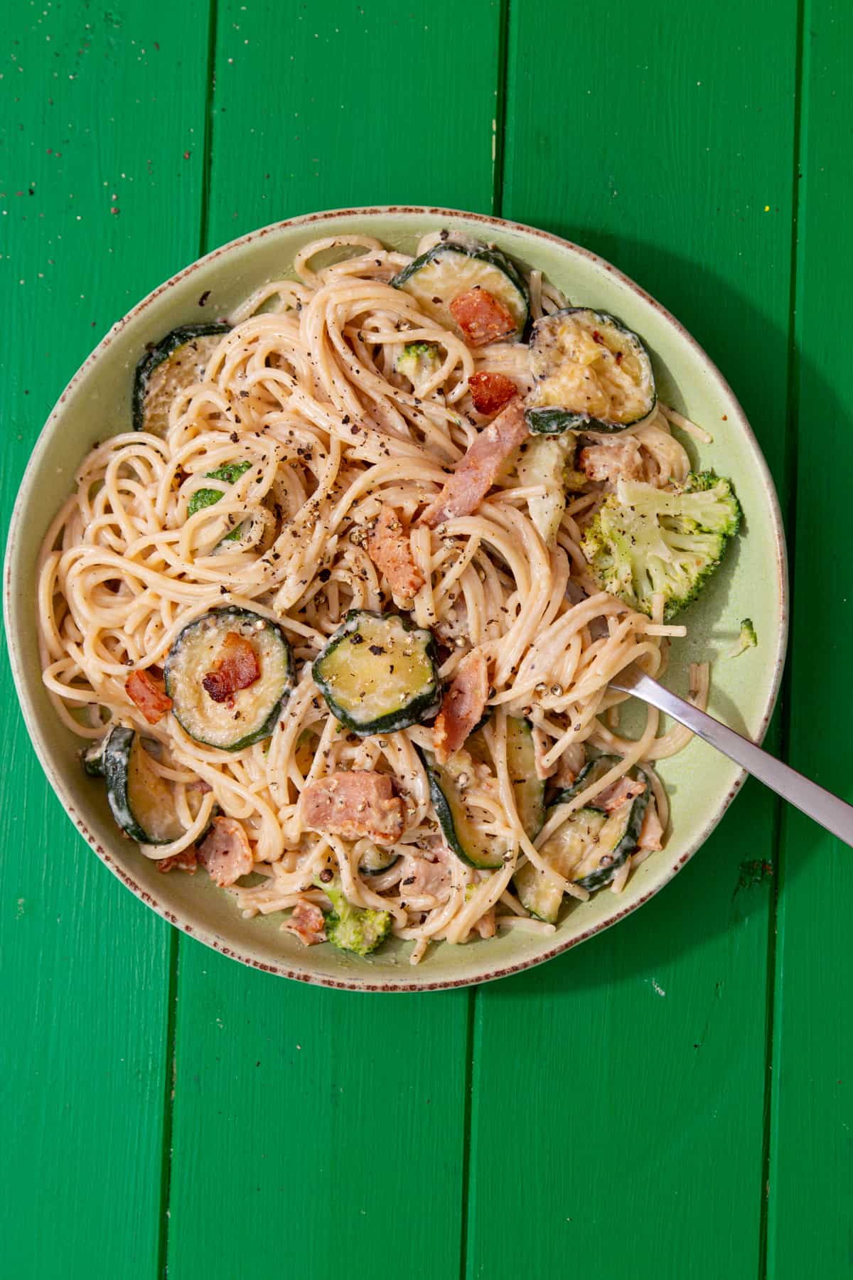 Creamy spaghetti with courgette rounds, bacon and broccoli in a bowl on a green background.