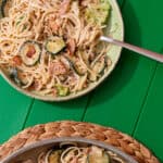 A bowl of creamy spaghetti, with bacon and courgettes with a spoon next to the spaghetti pan on a mat on a green background.