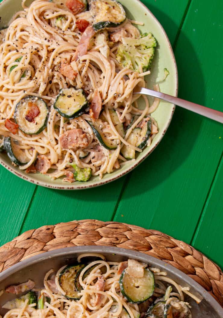 A bowl of creamy spaghetti, with bacon and courgettes with a spoon next to the spaghetti pan on a mat on a green background.