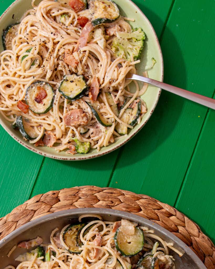 A bowl of creamy spaghetti, with bacon and courgettes with a spoon next to the spaghetti pan on a mat on a green background.