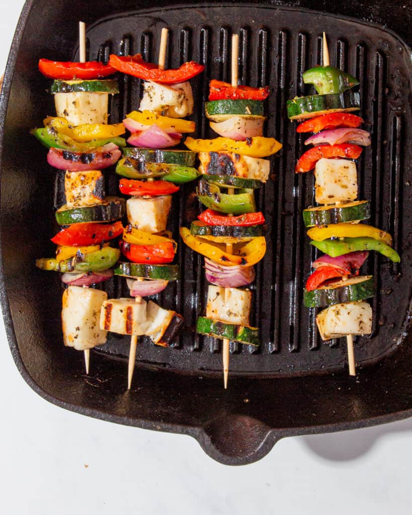 Halloumi and roasted vegetable kebabs on skewers on a griddle pan.