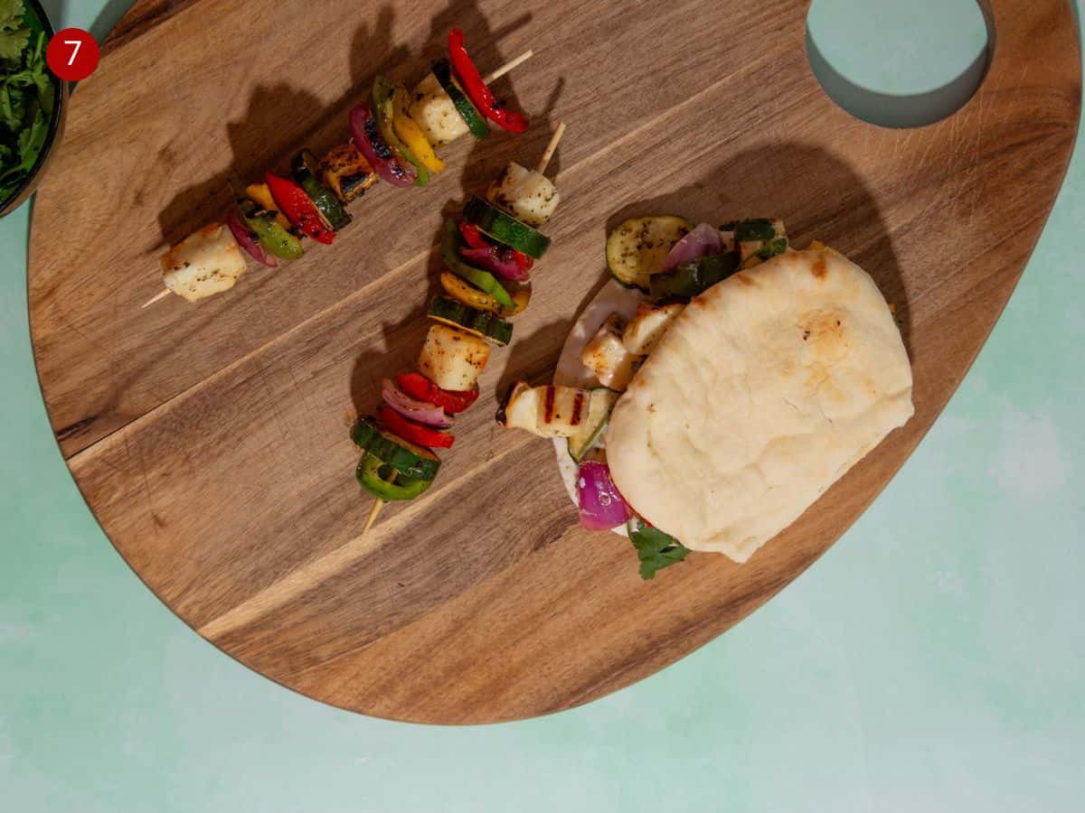 last process shot with 2 halloumi kebabs on sticks made with courgette slices, red onion and peppers and a filled flatbread with the roasted vegetables and halloumi inside on a wooden board.