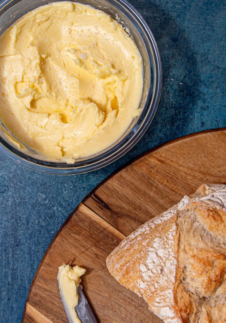 Butter in a round container next to a chopping board with a loaf of bread and knife with butter on it.
