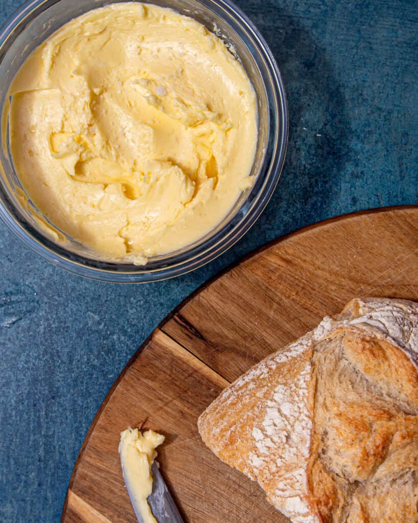 Butter in a round container next to a chopping board with a loaf of bread and knife with butter on it.