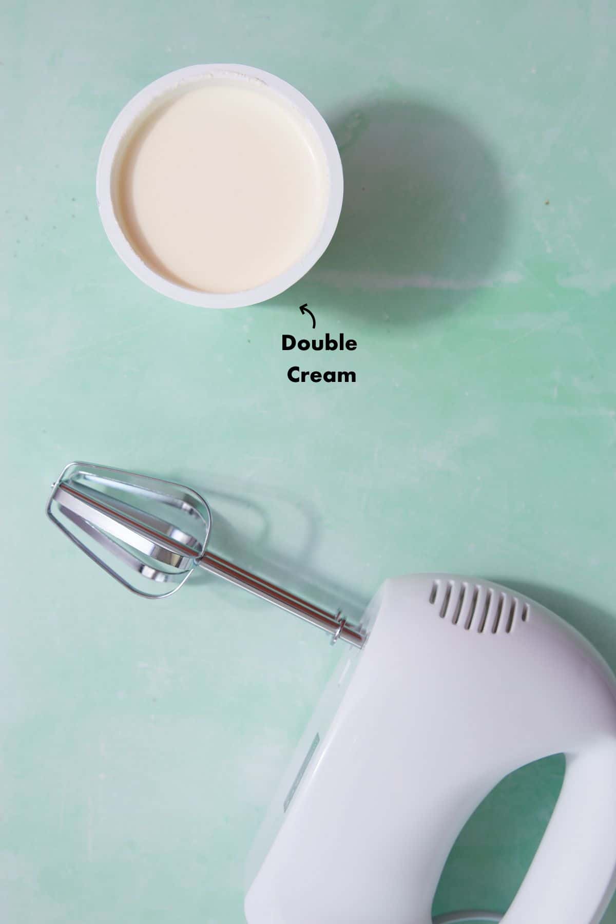 A pot of cream and a hand mixer laid out on a pale blue back ground.