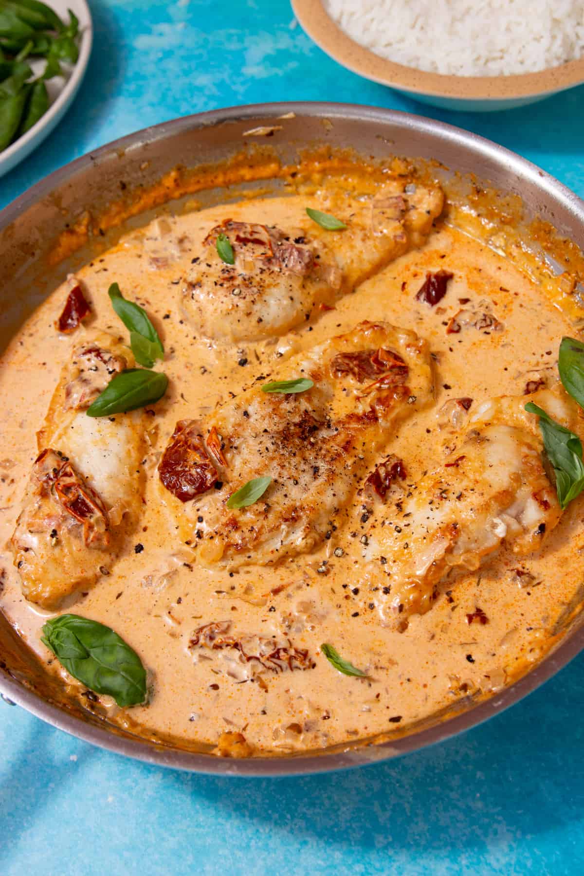 Chicken in a creamy tomatoey sauce in a large stainless steal pan topped with fresh basil on a turquiose blue background with a bowl of rice in background and basil in a bowl particially in view.