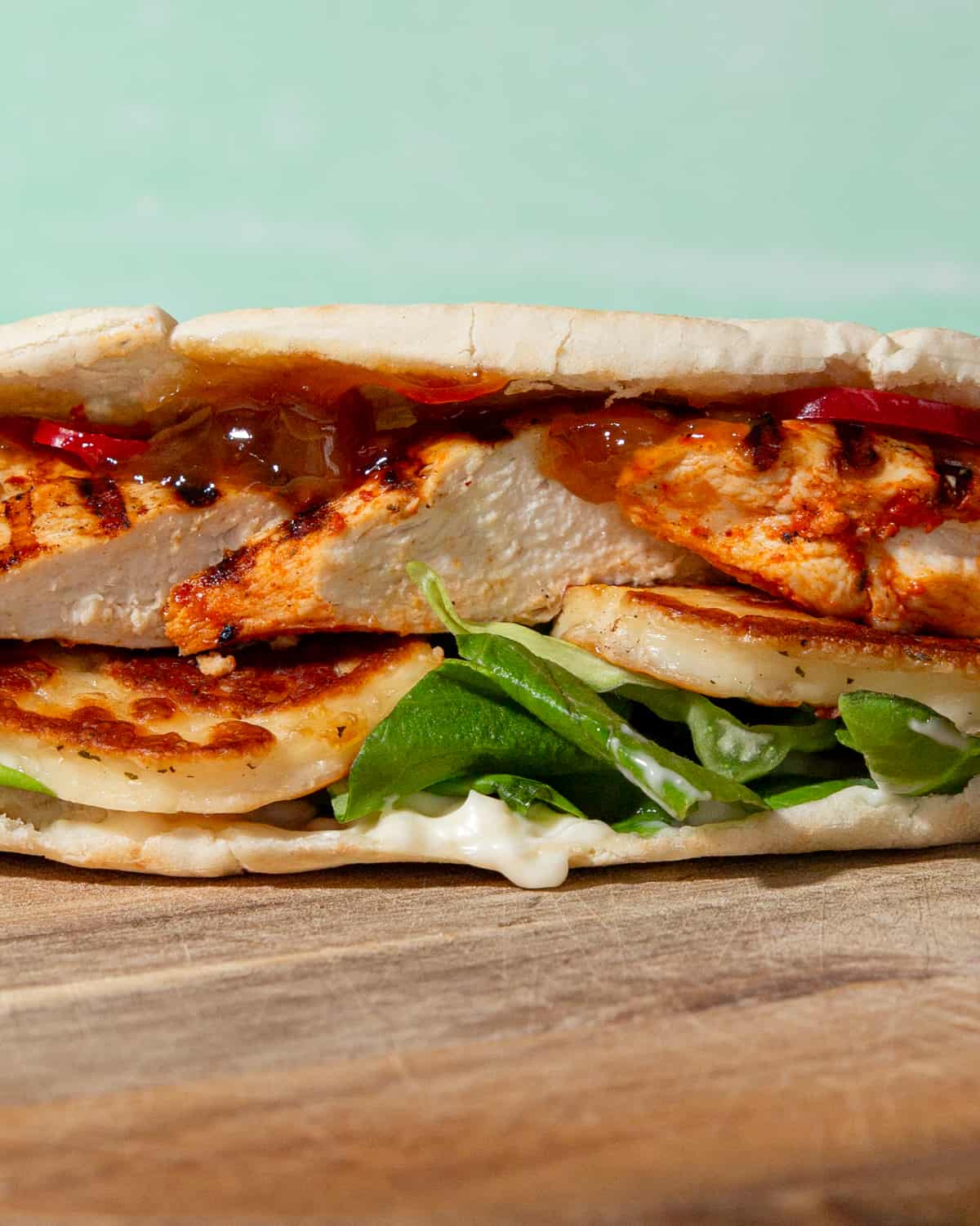 Side view of a white pitta filled with browned halloumi, lettuce, chicken, mayo and relish.