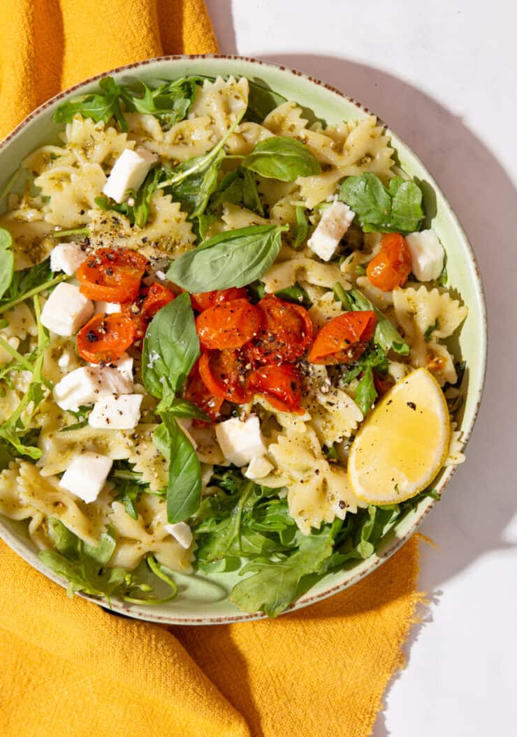 A bowl filled with pasta bows, topped with cherry tomatoes, basil leaves and a wedge of lemon on a mustard cloth.