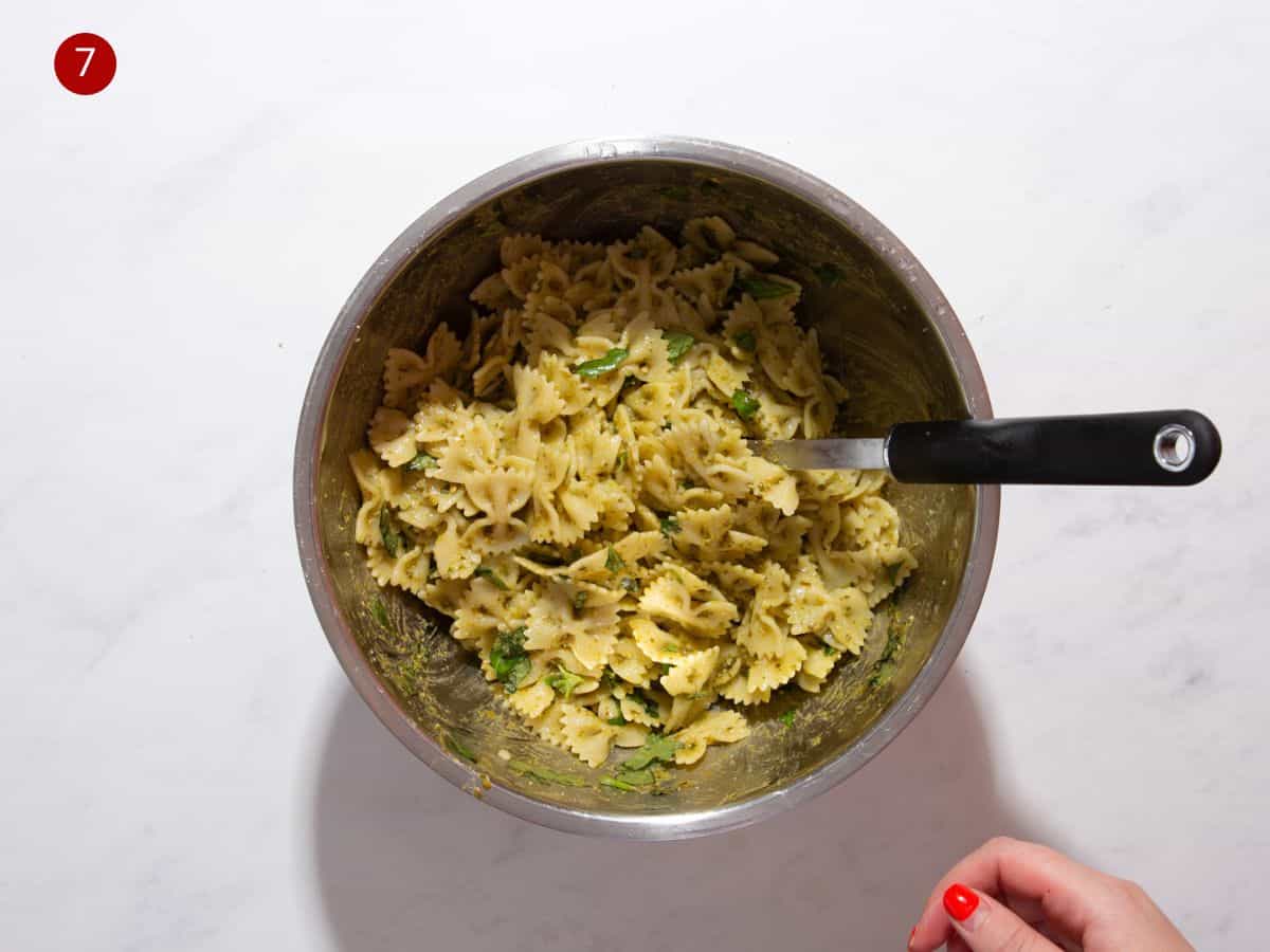 A large metal bowl with pasta bows mixed with basil and pesto with a spoon in the mix.