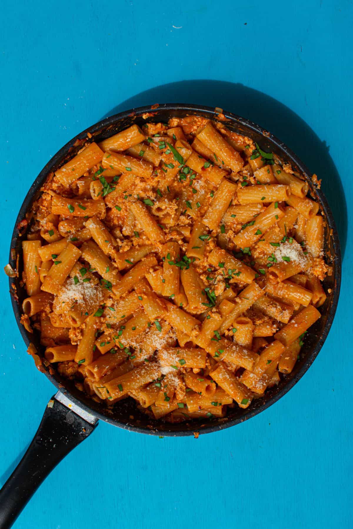 Overhead shot of rigatoni pasta with sauce topped with grated parmesan cheese and chopped basil in a large pan on a blue background.