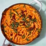 Overhead shot of pasta with a tomatoey sauce, sun dried tomatoes and sliced fresh basil in a large pan on a pale green background.