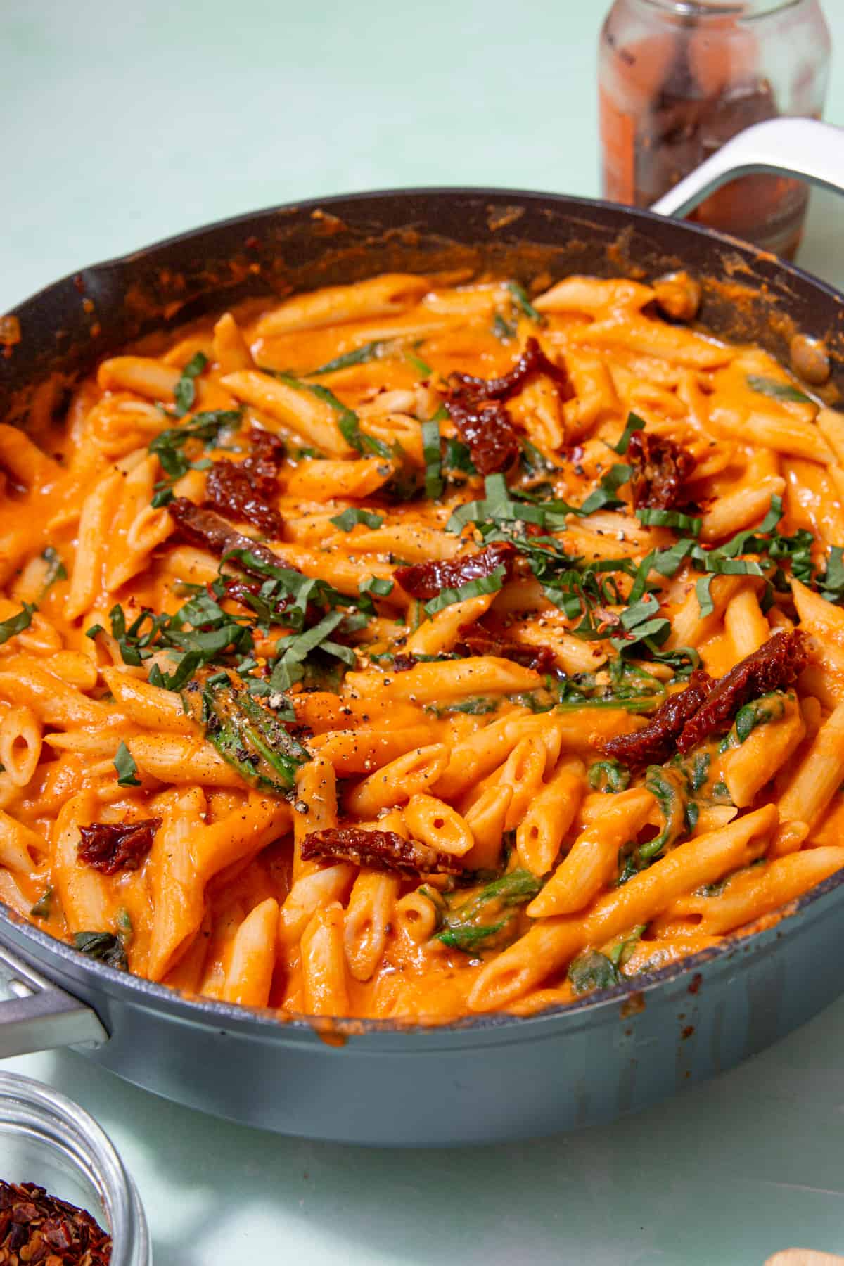 Close up of pasta with a tomatoey sauce, sun dried tomatoes and sliced fresh basil in a pan next to a jar of sun dried tomatoes and a small bowl infant of pan.