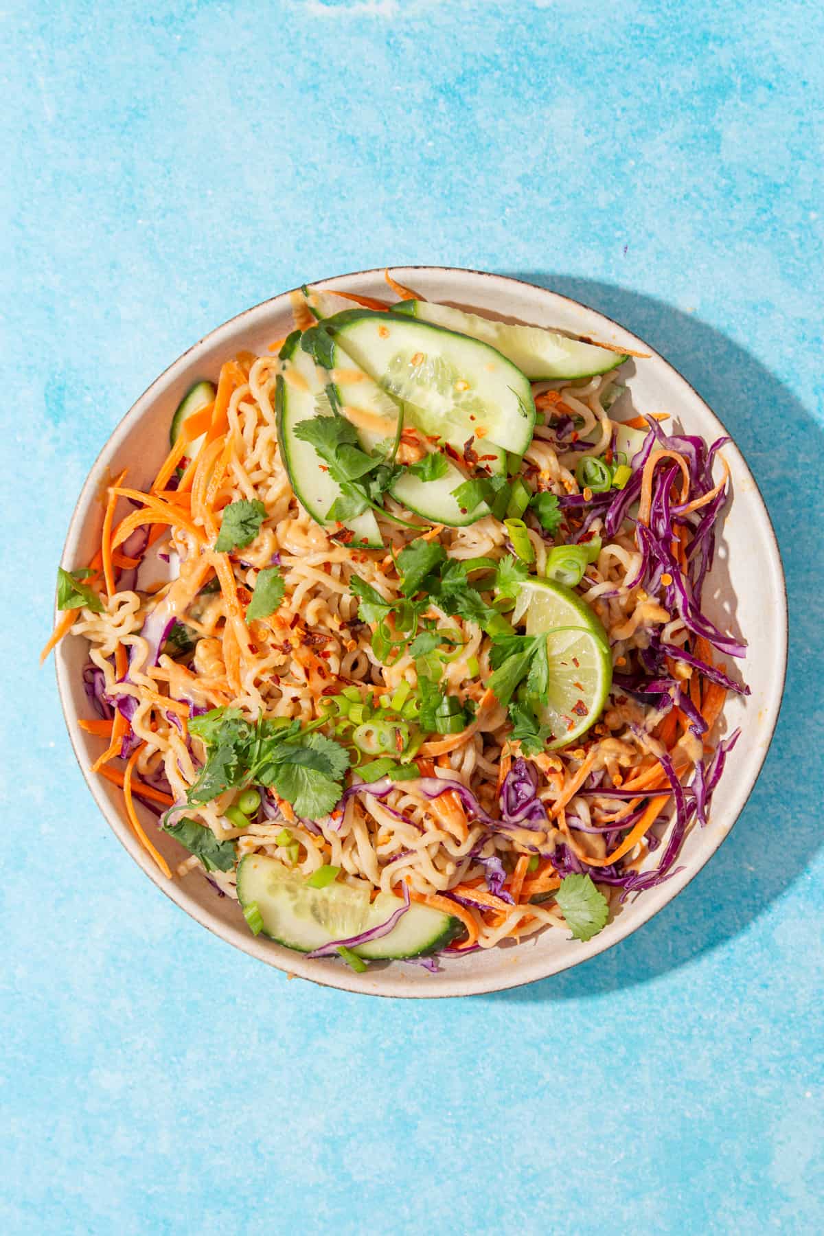 Overhead shot of a bowl of noodles, grated carrots, red cabbage, line wedges, spring onion and fresh coriander with peanut sauce on a blue background.