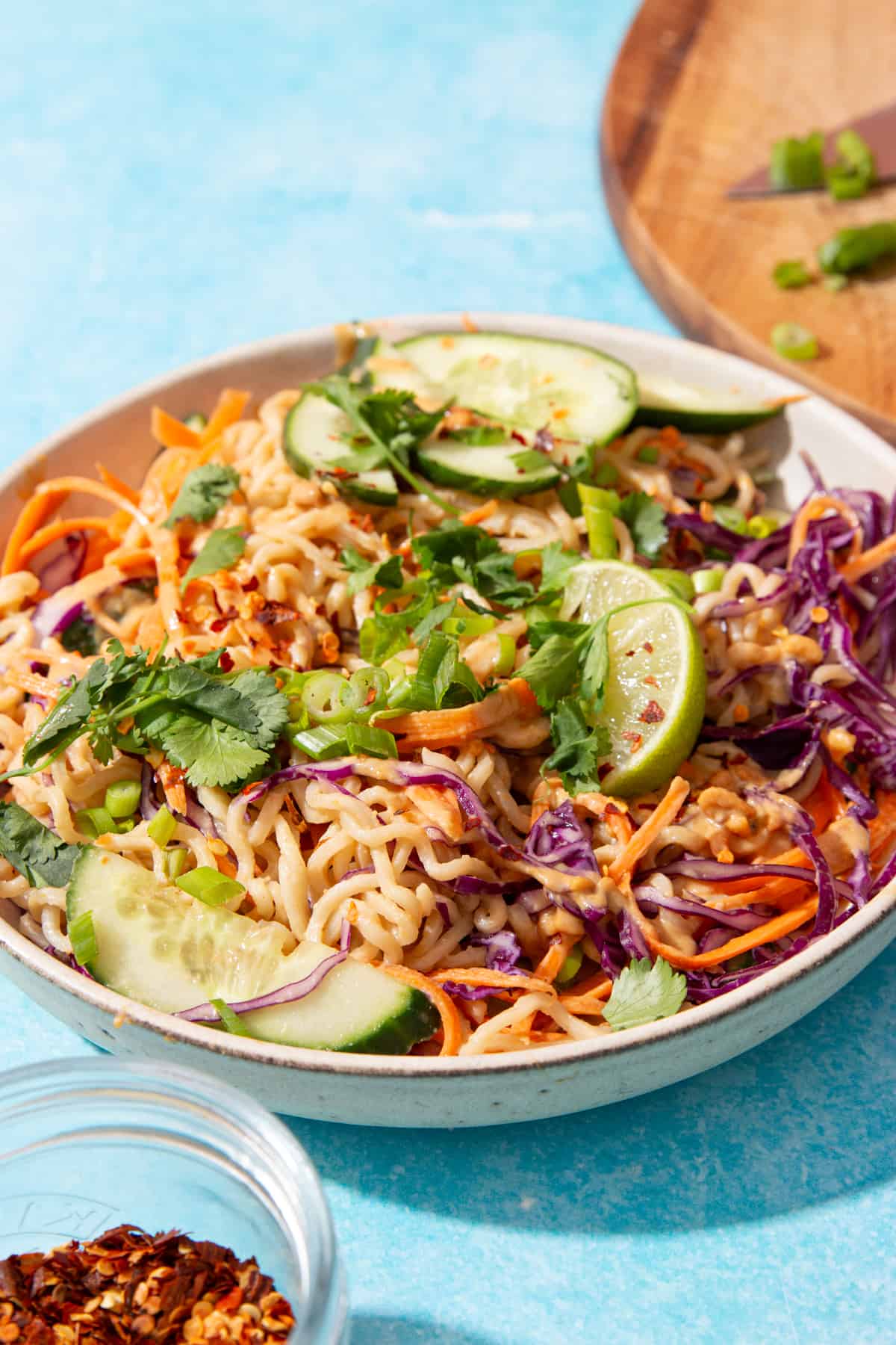 Size view of a bowl of noodles, grated carrots, red cabbage, line wedges, spring onion and fresh coriander with peanut sauce next to a small bowl of chilli flakes on a blue background..