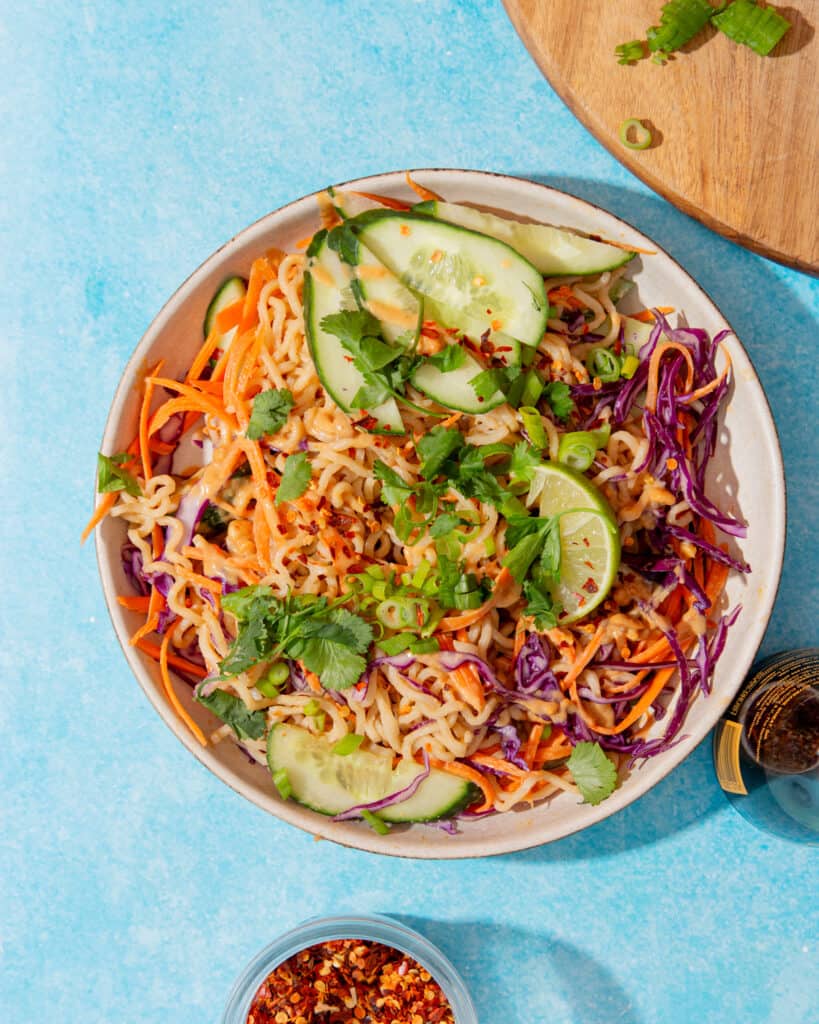 A bowl filled with shredded carrot, red cabbage, noodles cucumber and topped with coriander on a blue background.