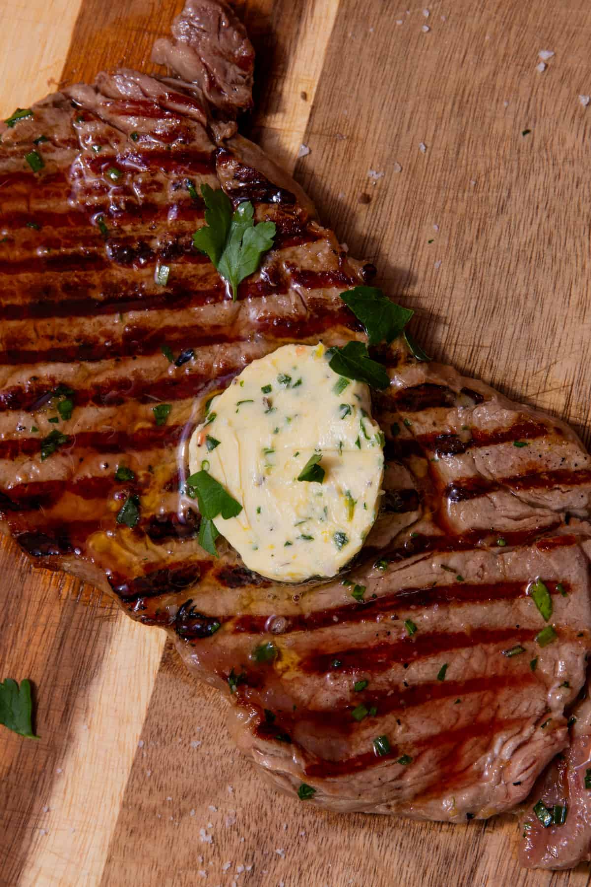 Close up a large piece of steak with char lines and a dollop of herby butter on a chopping board.