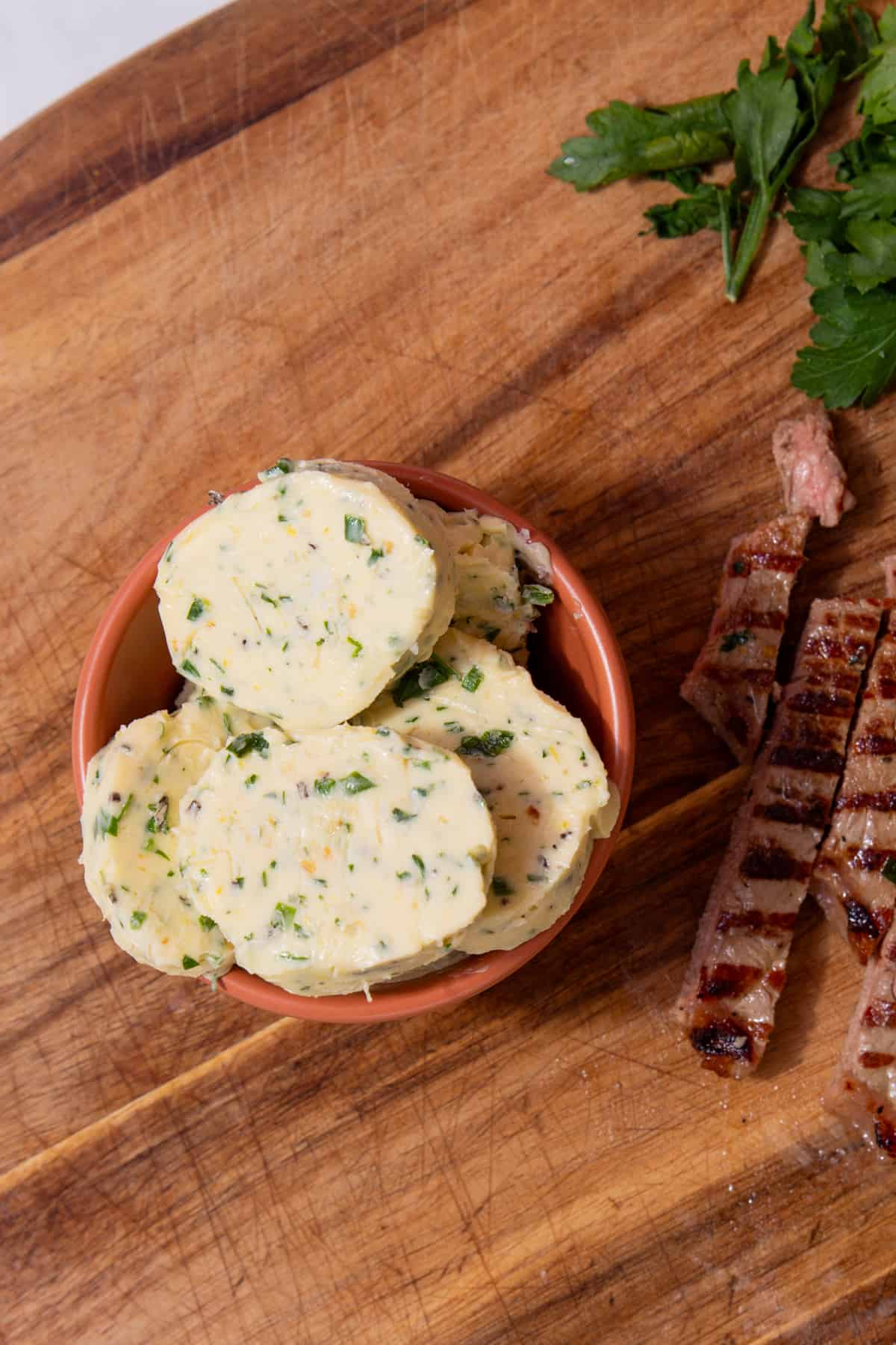 Rounds of herby butter in a smal bowl next to strips of charred steak on a wooden chopping board.