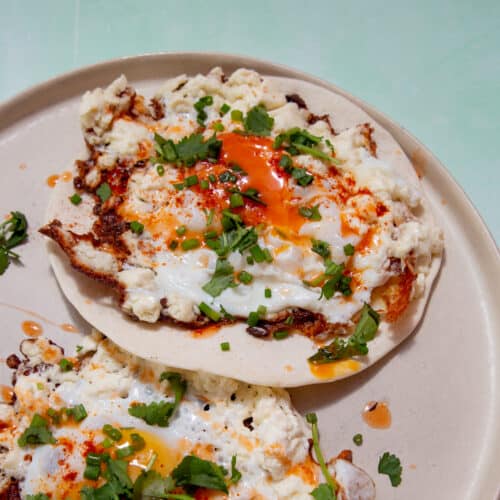 2 fried eggs on separate tortillas on a plate topped with coriander and chives on a pale green background.