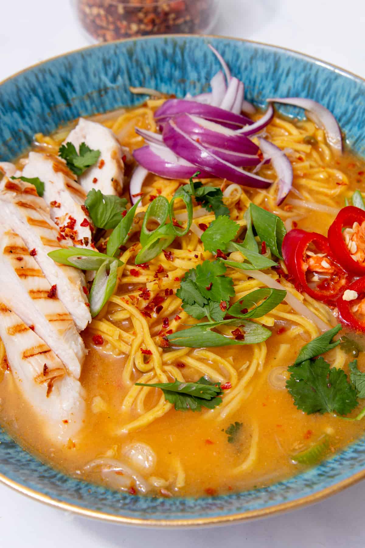 Close up of a blue bowl with slices of chicken, chillies, spring onions and red onion with noodles in soup next to a pot of chilli flakes.