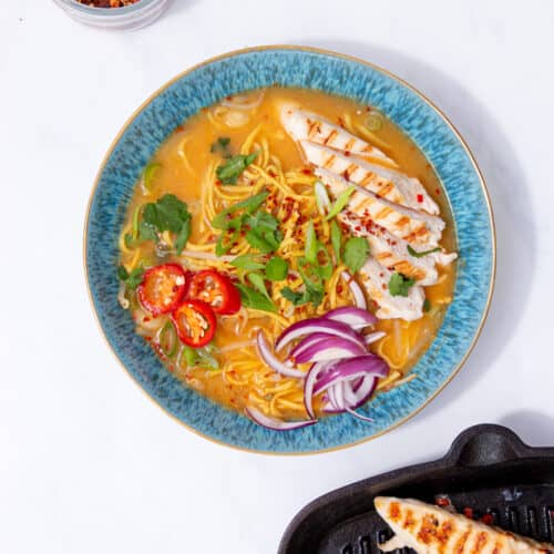 Overhead photo of a blue bowl with slices of chicken, chillies, spring onions and red onion with noodles in soup next to a pot of chilli flakes and a chopping board with extra chicken.