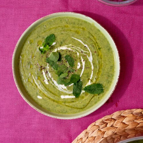 A bowl of courgette soup with a swirl of cream and garnished with fresh mint on a pink background next to the pan on a woven mat.