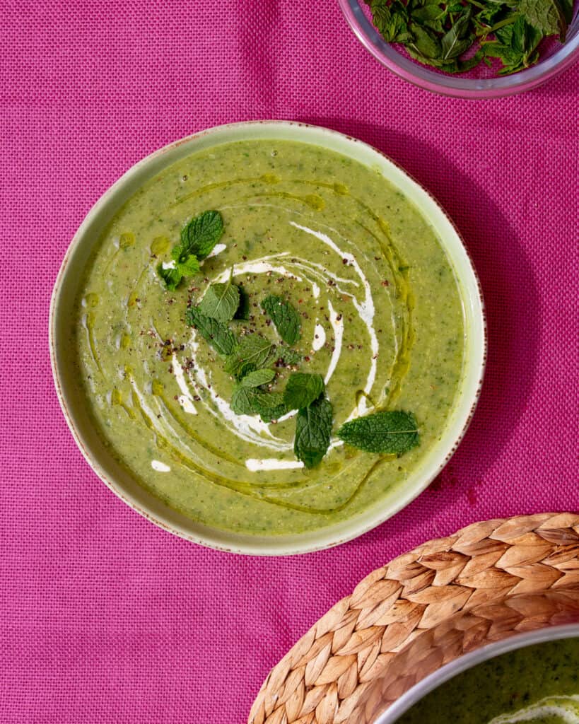 A bowl of courgette soup with a swirl of cream and garnished with fresh mint on a pink background next to the pan on a woven mat.