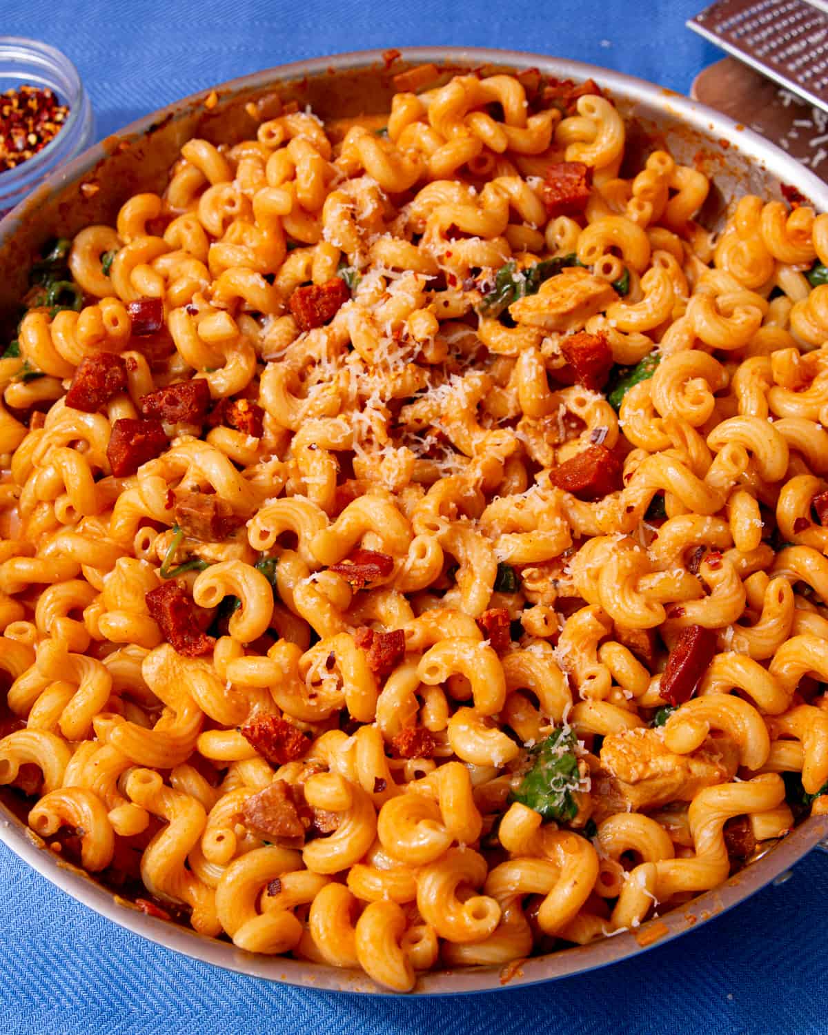 Close up of spirali pasta with bits of chorizo covered in a tomatoey sauce topped with some grated cheese in a stainless steel pan on a blue cloth.