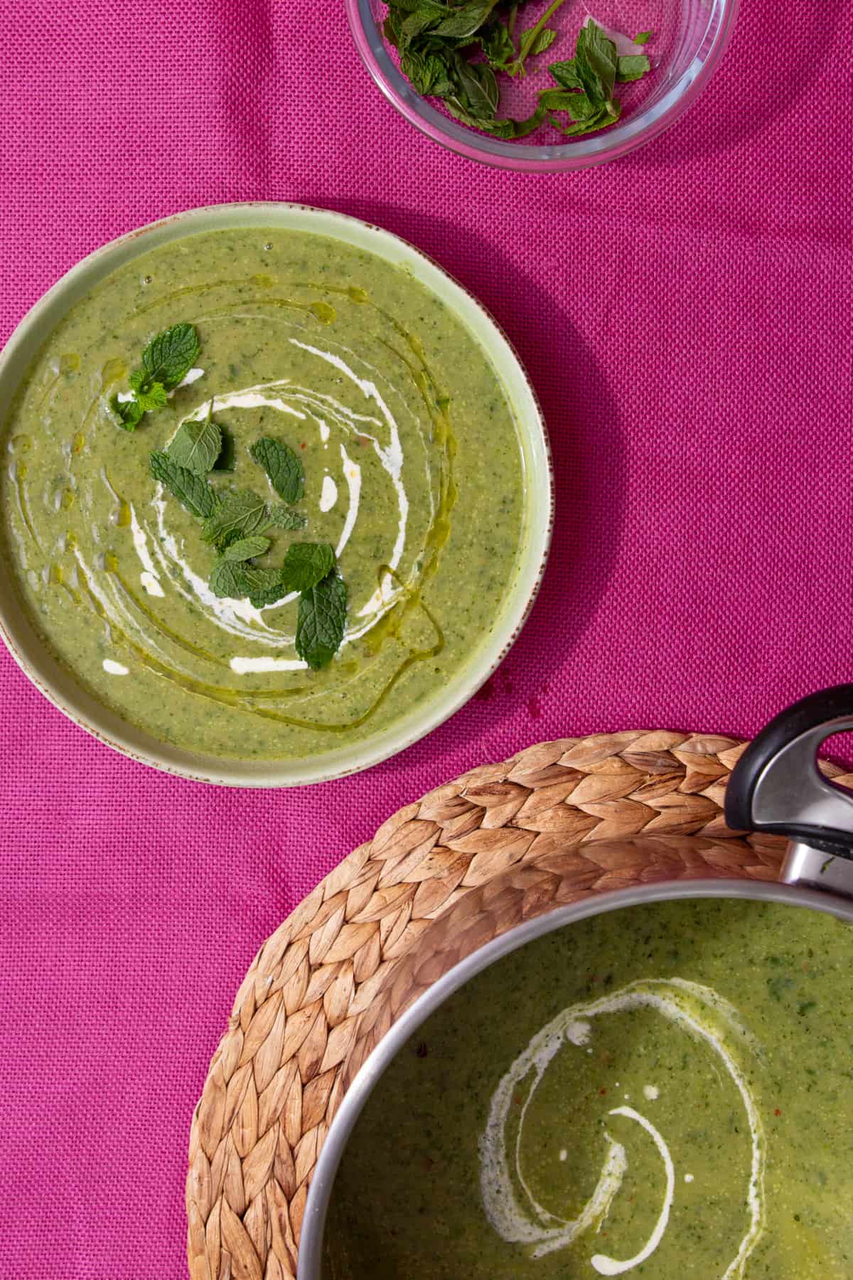 A bowl of courgette soup with a swirl of cream and topped with fresh mint leaves on a pink background with partial view of glass bowl with fresh mint and of the pan of soup on a woven mat.