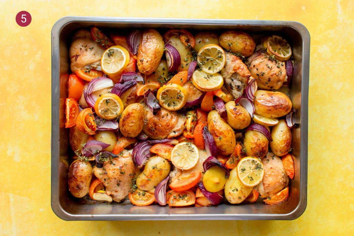 Overhead shot of golden browned chicken thighs, new potatoes and vegetables all baked with lemon rounds and red onions in a large baking tray on a yellow background.