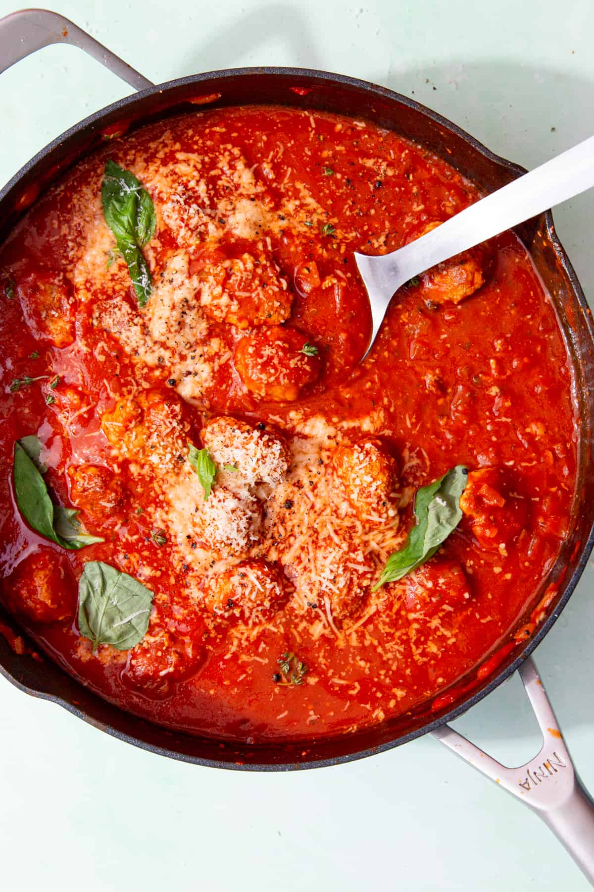 Meatballs in tomato sauce in a large pan garnished with fresh basil and some grated cheese with a serving spoon in the pan.