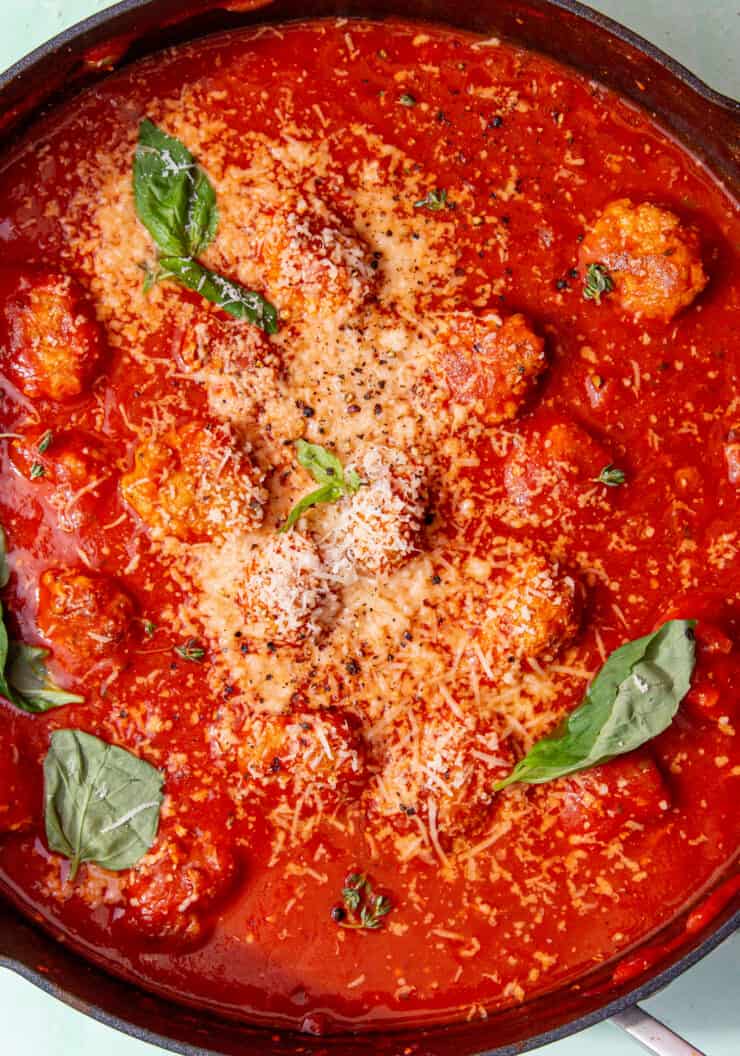 Meatballs in tomato sauce in a large pan topped with grated cheese and fresh basil leaves.