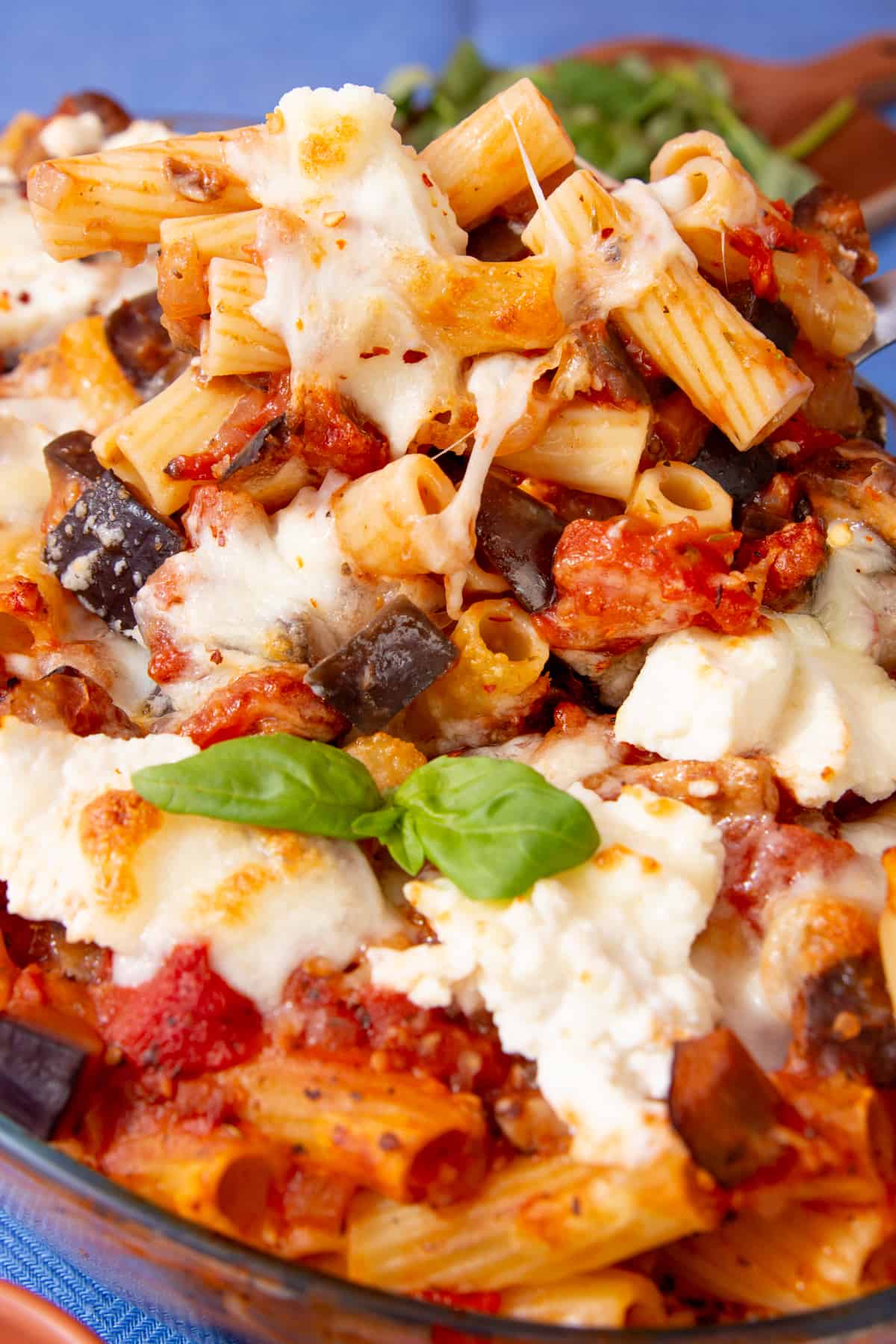 A close up of bowl of rigatoni pasta with a cheesy tomatey sauce with roasted vegetables and mozzarella cheese in a bowl with a wooden spoon in faint view.