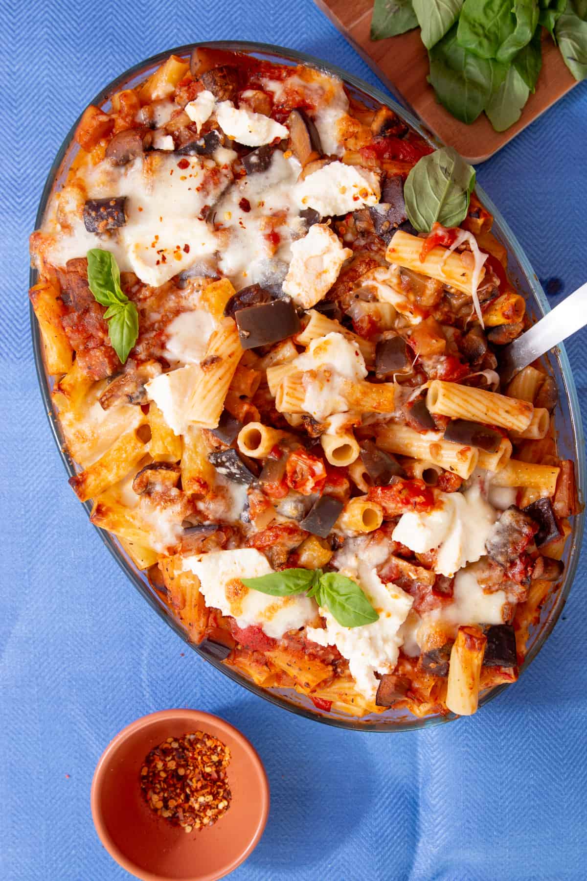 Overhead shot of of cooked rigatoni pasta with vegetables and melted cheese and tomato in an oval, glass bowl with fresh basil leaves as garnish and on a board behind the dish.