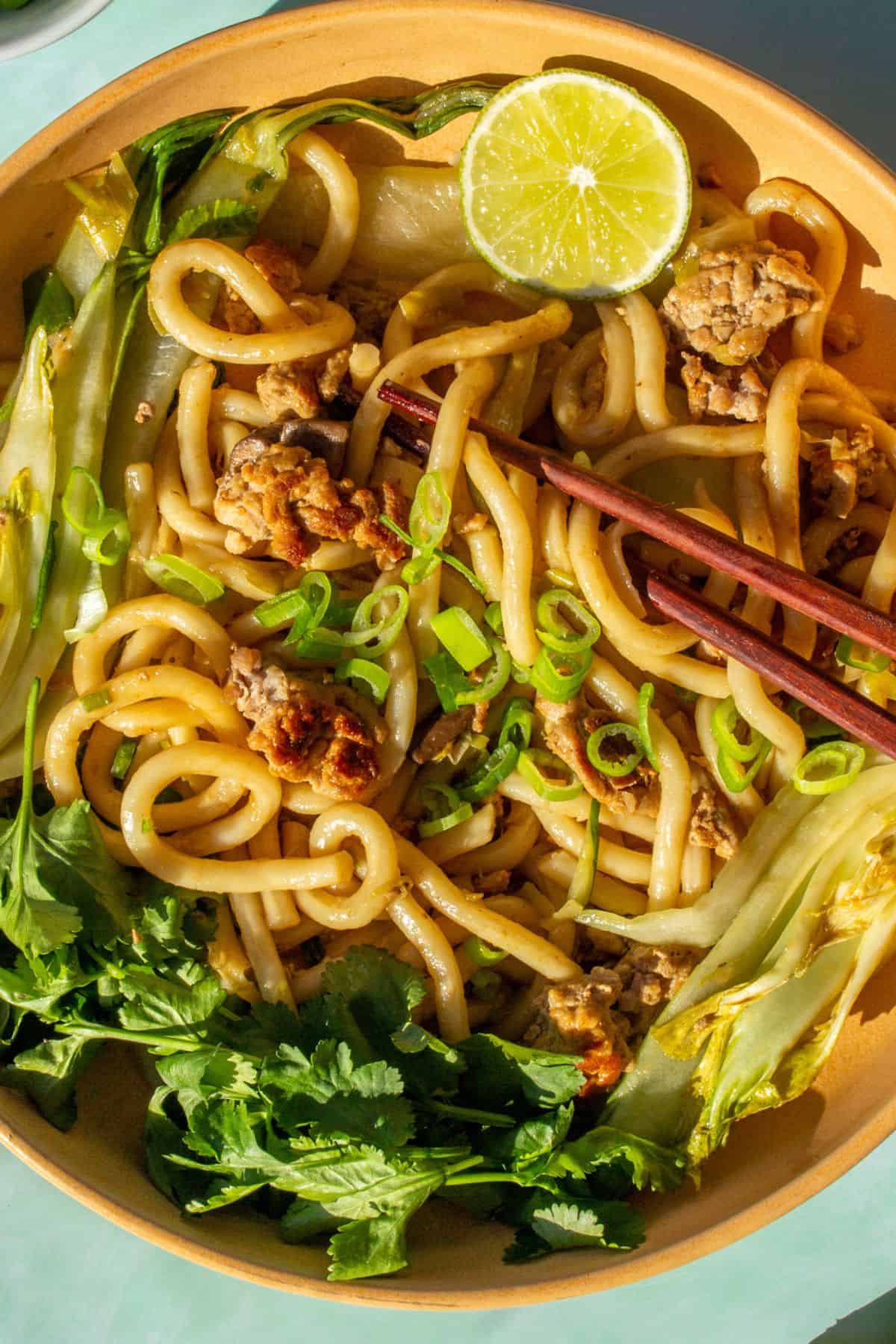 Close up of udon noodles in a bowl with browned mince, pok choi,sliced spring onion, a lime wedge and chop sticks.